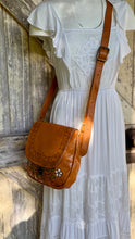 Load image into Gallery viewer, Woman wearing the Wildflower Bloom hand painted and hand tooled tan leather crossbody bag with floral artwork in a boho outdoor setting.
